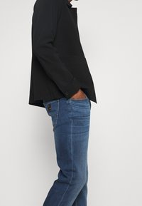 Man med svart långärmad tröja och händerna i fickorna, samt blå jeans, framför en ljus enfärgad bakgrund.