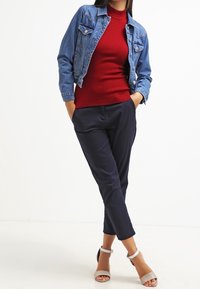 Vrouw die met handen in haar zakken staat, gekleed in een blauwe spijkerjack, een rode trui, marineblauwe broek en beige haksandalen.