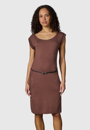 Frau mit lockigem Haar, die ein braunes knielanges Kleid mit kurzen Ärmeln und einem dünnen schwarzen Gürtel trägt, steht vor einem schlichten Hintergrund.