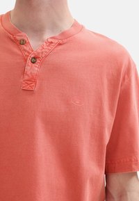 Korálové bavlnené tričko s Henley výstrihom, s dvoma gombíkmi na zapínaní a malým logom na ľavej hrudi, hladká textura a krátke rukávy.