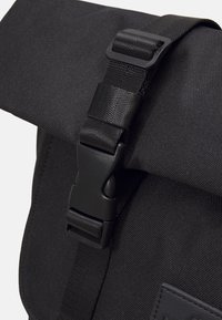 Musta olkalaukku, jossa on teksturoidusta kankaasta valmistettu pinta, taitettava läppä ja musta muovinen soljella varustettu kiinnitys turvallista sulkemista varten.