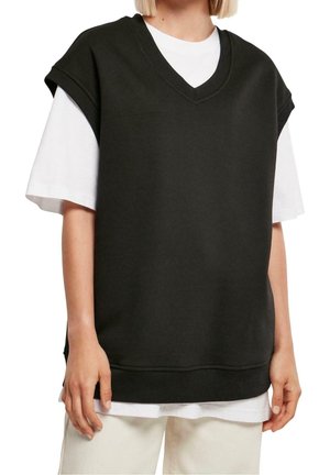 Person trägt einen schwarzen ärmellosen V-Ausschnitt-Pullover über einem weißen Kurzarm-T-Shirt und hellen Hosen, steht mit entspannten Händen.