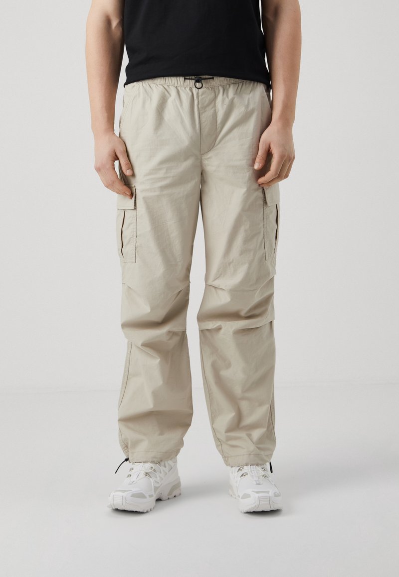 Personne portant un pantalon cargo beige avec plusieurs poches et des chaussures de sport blanches, debout devant un fond clair uni.