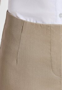 Beige stoffen broek met een zachte textuur, subtiele glans, rechte snit en gestikte zakken op de taille. Witte shirt zichtbaar aan de bovenkant.