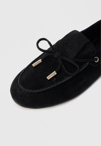 Mustad seemisnahast mokassiinid ümarate ninaosadega, millel on dekoratiivne lips ja metallist detailid. Pind on sileda tekstuuriga ja õmblused on peened.