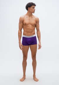 Νεαρός μυώδης άνδρας που στέκεται ξυπόλητος με μωβ εσώρουχα Polo Ralph Lauren, μπροστά σε απλό ανοιχτό γκρι φόντο, κοιτάζοντας προς τα δεξιά.