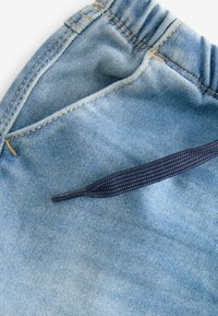 Tissu denim bleu clair avec une surface texturée, doté d'un cordon de serrage bleu marine et de coutures contrastantes le long des coutures et des poches.