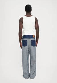 Un homme de dos portant un t-shirt sans manches blanc et un jean ample avec des poches en denim foncé et une ceinture assortie, sur un fond uni.