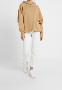 Beżowa, za duża bluza z kapturem, z kieszenią typu kangur i czarnym logo na piersi, zestawiona z białymi prostymi spodniami i białymi trampkami.