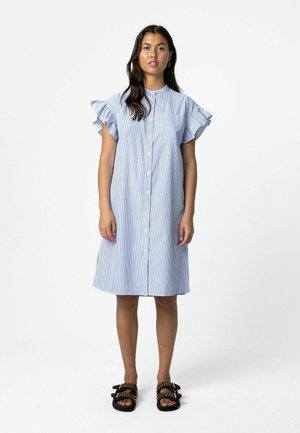 Žena stojaca v svetlomodrých krátkych šatách na gombíky s volánovými rukávmi a čiernymi sandálmi s cvokmi na jednoduchom bielom pozadí.