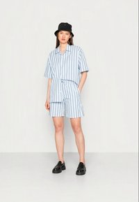 Camisa de manga curta riscada em azul-claro combinada com calções à medida. Apresenta um corte relaxado, riscas verticais e sapatos grossos pretos.