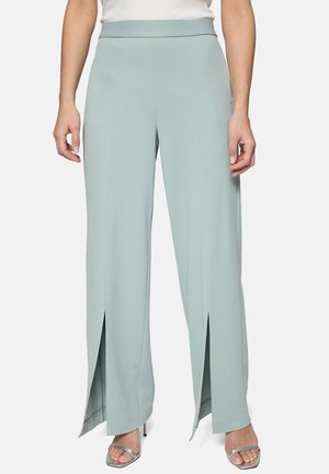 Femme portant un pantalon large vert clair avec des fentes à l'avant de l'ourlet, assorti à des talons ouverts argentés et un haut blanc.
