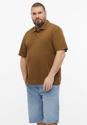 Mann mit Bart und zurückgebundenem Haar, der ein braunes kurzärmeliges Poloshirt und hellblaue Jeansshorts trägt, stehend vor einem schlichten weißen Hintergrund.