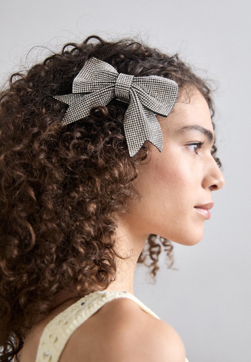 Srebrny klips do włosów w kształcie wstążki obszyty błyszczącymi kamieniami, o teksturowanym wykończeniu i szerokim, zakręconym kształcie, umocowany w kręconych włosach.