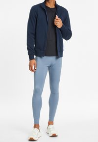 Chaqueta navy con cremallera, camiseta negra y leggins deportivos de color azul claro. Zapatillas blancas tipo slip-on con un sutil patrón de camuflaje. Texturas de tela suaves.