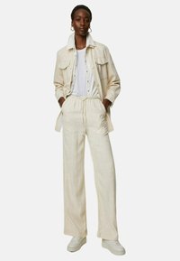 Ljusbeige jacka med stora fickor, i kombination med matchande beige löst sittande byxor och en vit t-shirt. Sneakerskompatibla skor fulländar looken.