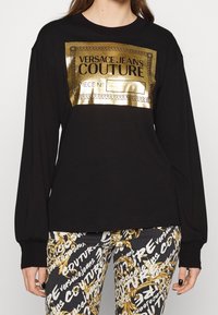 Camiseta negra de manga larga con un estampado metálico dorado del logo "VERSACE JEANS COUTURE" y un borde decorativo. Acompañada de pantalones estampados.