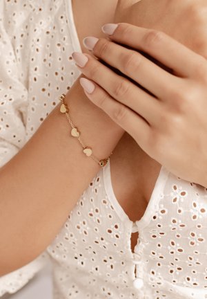 Goldarmband mit kleinen herzförmigen Anhängern und einer zarten Kette, getragen an einem Handgelenk mit gepflegten Fingernägeln. Weiße bestickte Bluse.