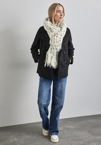 Sort dobbeltradet frakke med tekstureret stof, cremet tørklæde med sorte prikker, blå jeans og hvide sneakers, sat mod en neutral baggrund.