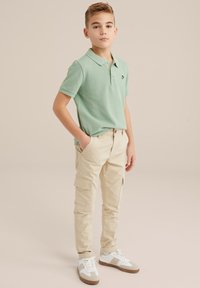 Polo majica iz svetlozelene tkanine sa kratkim rukavima, uparena sa bež cargo pantalonama koje imaju bočne džepove, i belim patikama sa bež detaljima.