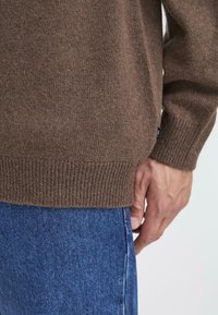 Smeđi pleteni pulover s rebrastim rubom, teksturiranom tkaninom i bočnim prorezima, uparen s plavim trapericama. Ruka počiva na struku.