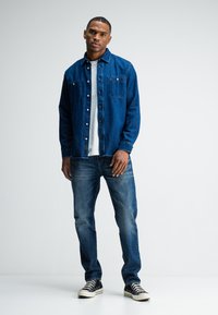 Denim overhemd in donkerblauw, met knopen, twee borstzakken, gecombineerd met vervaagde blauwe jeans en zwarte sneakers met witte zolen.
