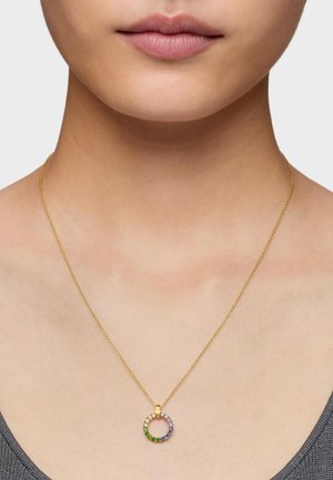 Primer plano de una persona que lleva un collar de cadena dorada con un colgante circular que presenta piedras preciosas multicolores.