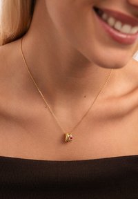 Primo piano di una donna che indossa una delicata collana d'oro con tre piccoli anelli d'oro, uno dei quali presenta accenti di gemme rosse e bianche.