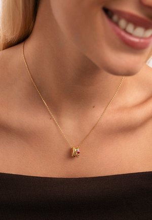 Primer plano de una mujer que lleva un delicado collar de cadena de oro con tres pequeños anillos de oro, uno de los cuales presenta acentos de piedras preciosas rojas y blancas.