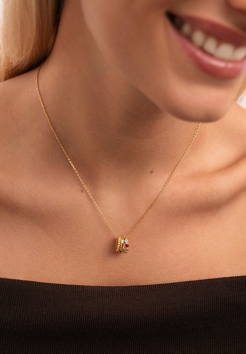 Primer plano de una mujer que lleva un delicado collar de cadena de oro con tres pequeños anillos de oro, uno de los cuales presenta acentos de piedras preciosas rojas y blancas.