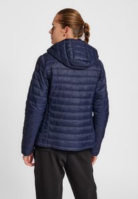 Marineblauwe pufferjack met verticale stiksels, een capuchon en een gladde, glanzende textuur. Voorzien van ritsluiting en elastische manchetten.