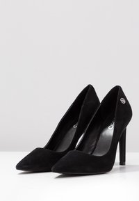 Zapatos de tacón alto de ante negro con punta afilada, diseño elegante y logotipos en el lado exterior. Forro interior brillante y tacones de aguja.