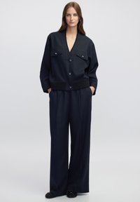 Chaqueta azul marino con dos bolsillos frontales y escote en forma de V, combinada con pantalones anchos azul marino. Ambos tienen una textura suave y un diseño minimalista.