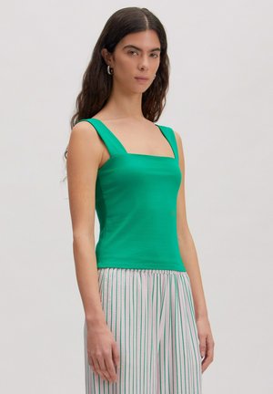 Jeune femme aux longs cheveux foncés portant un haut sans manches vert et un pantalon blanc à rayures verticales vertes, debout devant un fond uni.