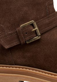 Bottines en suède marron avec une boucle décorative en laiton, des détails cousus et une semelle texturée pour une meilleure adhérence.