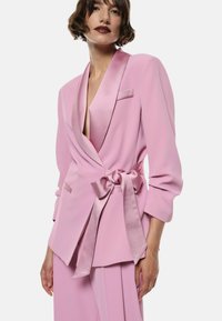 Lys pink blazer med satin revers og bælte. Har lange ærmer, en dyb V-hals og en frontal lomme. Glat tekstur.