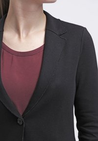 Czarna marynarka z ostrym klapem i jednym guzikiem, nałożona na bordową bluzkę, wykonaną z gładkiego materiału i o dopasowanym kroju.