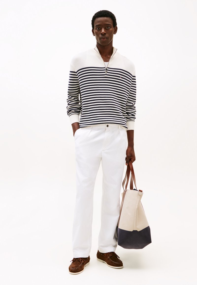 Suéter de cuello con cremallera a rayas en azul marino y blanco, pantalones blancos, zapatos de barco marrones y un bolso de dos tonos con un diseño en beige y azul marino oscuro.