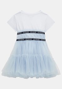 Biała bluzka z krótkim rękawem przymocowana do jasnoniebieskiej spódnicy z tiulu, z czarnymi pasami marki "GUESS" na talii i biodrach, widok z tyłu.