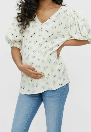 Gelbe blumige Umstandsbluse mit Puffärmel, V-Ausschnitt und lockerer Passform. Kombiniert mit hellblauen Jeans. Zarte Textur und Muster.