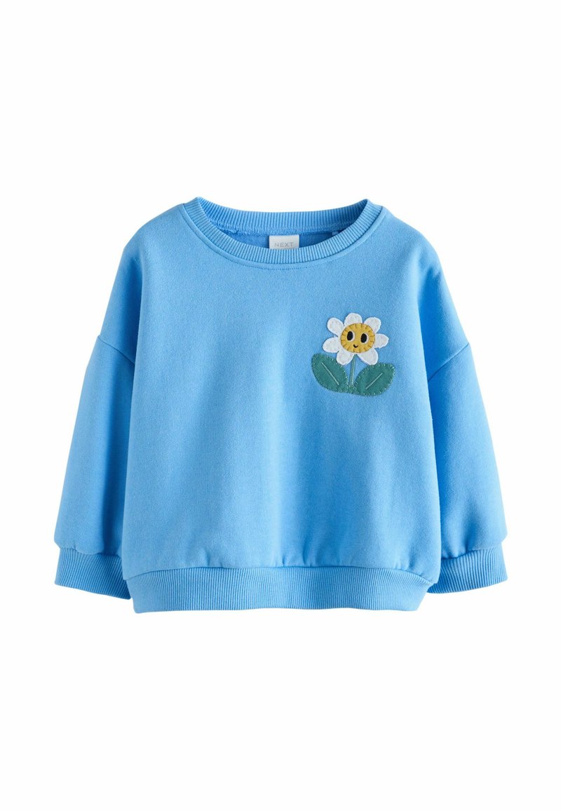 Világoskék pulóver hosszú ujjakkal, kerek nyakkivágással, és a mellrészen sárga közepű, zöld levelekkel díszített virágalkalmazással.