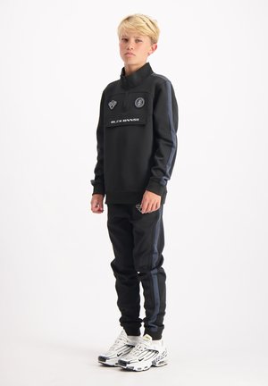 Blond meisje dat in een zwart trainingspak staat met grijze zijkanten en witte sneakers op een witte achtergrond, iets naar links gekeerd.