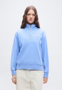 Νεαρή γυναίκα με μακριά καστανά μαλλιά, φορώντας ένα ανοιχτό μπλε φούτερ με φερμουάρ μέχρι τη μέση και εκρού παντελόνι, στέκεται μπροστά σε απλό φόντο.