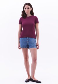 Maglietta bordeaux a maniche corte abbinata a pantaloni corti in denim azzurro. Modella che indossa scarpe basse nere, con sfondo neutro. Design semplice.