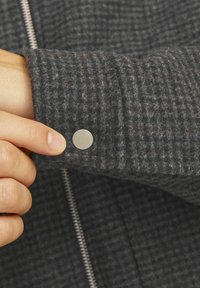 Manche de veste en tissu texturé gris avec un bouton-pression métallique, montrant une main tenant la manche près d'une fermeture éclair.