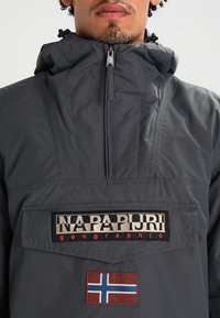 Mann trägt eine dunkelgraue, mit Reißverschluss versehene Jacke mit dem Aufnäher "NAPAPIJRI geographic" und einem kleinen norwegischen Flaggenaufnäher auf der Vorderseite.