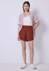 Pantaloni corti in lino color ruggine con una cintura elastica, abbinati a una canotta bianca e a una camicia a righe, leggermente svasata e con bottoni.