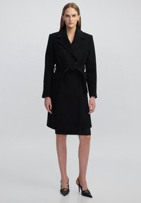 Cappotto di lana nero con design doppiopetto, grande colletto, vita cintrata con cintura e tasche laterali, abbinato a una gonna a tubino nera e tacchi a punta.