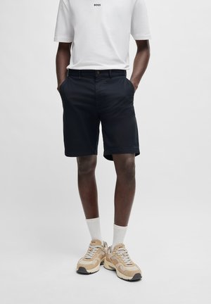 Shorts navy à coupe décontractée, fabriqués en tissu léger, avec des poches latérales et une fermeture à bouton, associés à des baskets beiges.