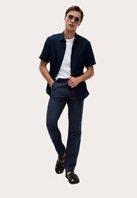 Korte mouwen, marineblauw overhemd met knopen over een wit t-shirt, gecombineerd met een donkere broek en zwarte sandalen. Model staat met handen in de zakken.